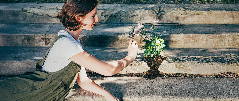 Mädchen schaut mit Lupe eine Pflanze an die aus Beton wächst