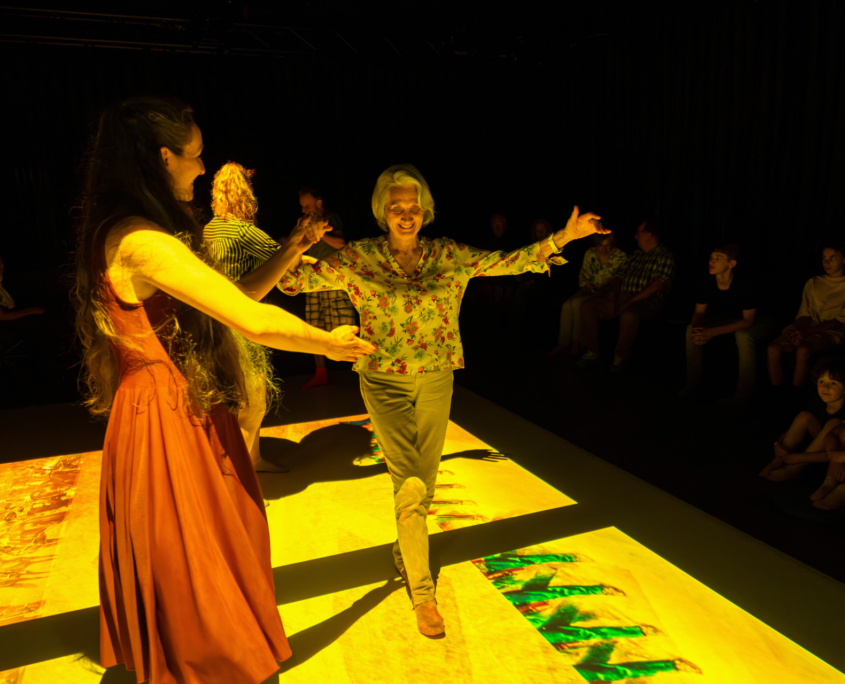 Eine Bühne mit Projektionen. Auf der Bühne tanzt eine Tänzerin mit einer älteren Besucherin. Im Hintergrund sind weitere Besucher*innen. Neben der Bühne blicken Zuschauer*innen auf die Bühne.